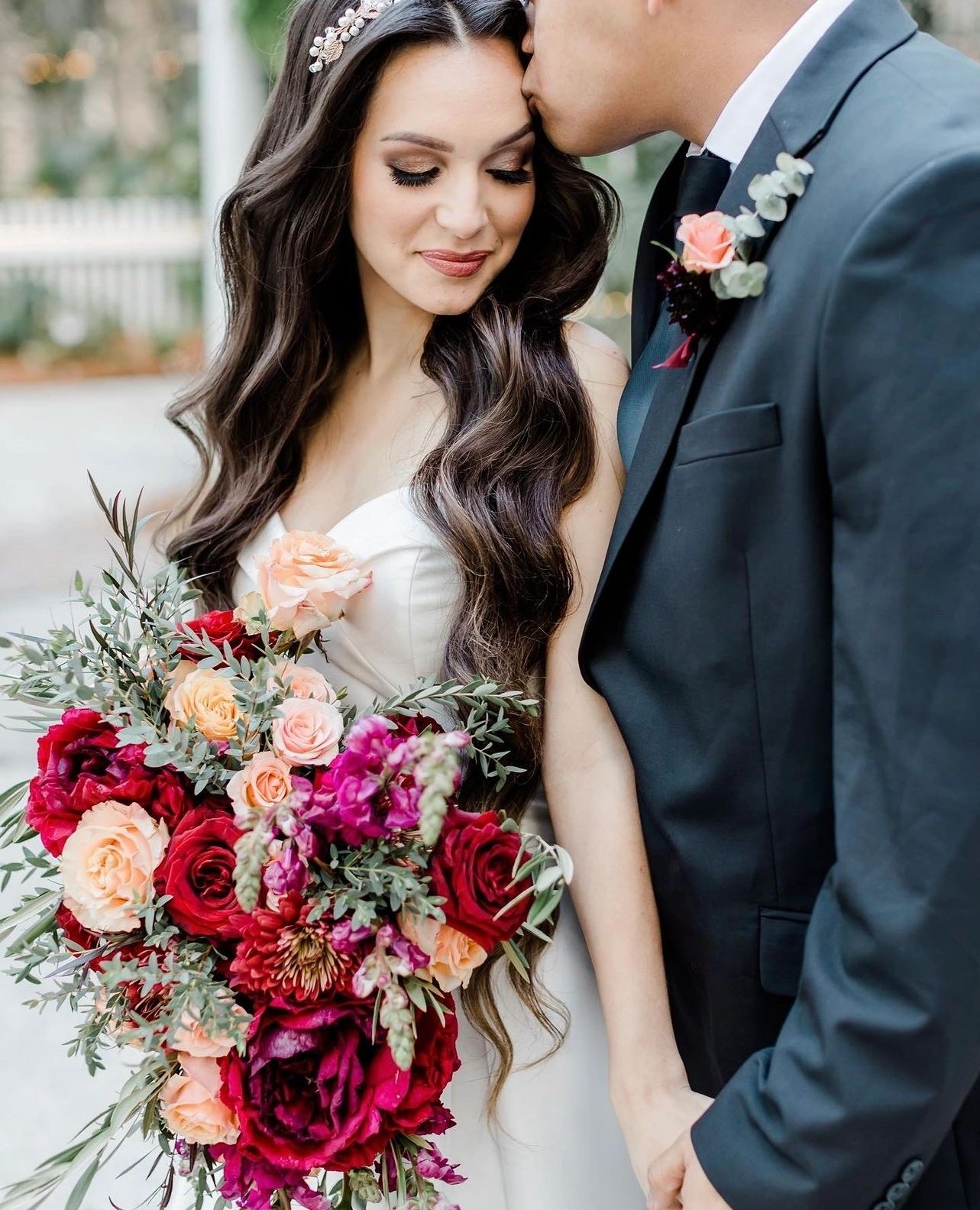 Groom kissing bride's forehead, bride holding vibrant rose bouquet